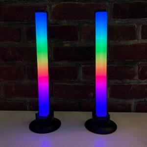 Due barre luminose LED a tubo verticali, che mostrano un gradiente di colori dal blu al viola, su basi nere, davanti a un muro di mattoni.