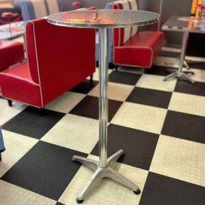 Tavolo alto da bar in metallo lucido con piano circolare testurizzato e base a quattro razze, in un ambiente stile diner.