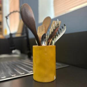 Portautensili cilindrico giallo in ceramica con cucchiai in legno e utensili in metallo, su un piano di lavoro scuro.