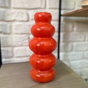 Vaso decorativo in vetro arancione brillante con forma a lobi sovrapposti, posizionato su una mensola di legno.