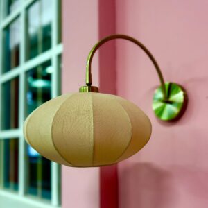 Applique da parete con paralume ovoidale in tessuto beige e braccio curvo in metallo dorato, montata su una parete rosa.