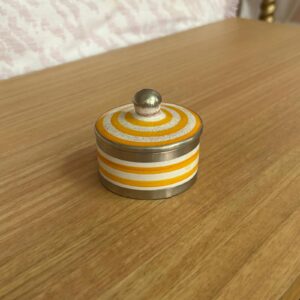 Piccola scatola decorativa rotonda con coperchio, base in metallo argentato e corpo a strisce orizzontali bianche e arancioni. Il coperchio ha cerchi concentrici e un pomello sferico.