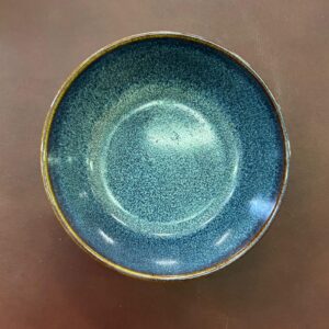 Ciotola in ceramica con smalto reattivo blu-verde e texture granulosa, vista dall'alto su sfondo marrone.