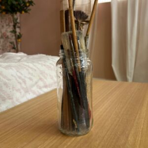 Vaso in vetro trasparente riempito con numerosi pennelli da pittura di varie dimensioni e usura, su un tavolo di legno chiaro.