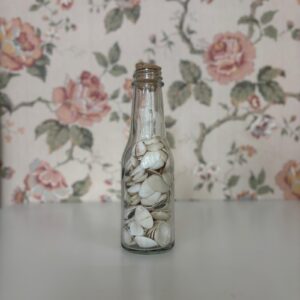 Bottiglia di vetro trasparente riempita di piccole conchiglie bianche e beige, con tappo di sughero, su sfondo floreale.