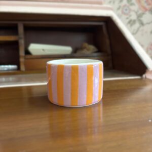 Contenitore cilindrico in ceramica con strisce verticali rosa lavanda e arancioni, posizionato su una superficie di legno.