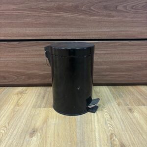 Cestino a pedale cilindrico di colore nero opaco, posizionato su un pavimento in legno chiaro davanti a un mobile in legno scuro.