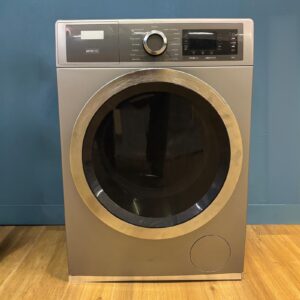 Lavatrice moderna grigia con oblò metallico e pannello di controllo digitale, posizionata su pavimento in legno davanti a parete blu.