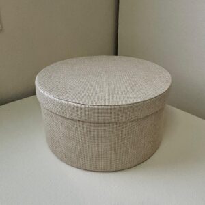 Cofanetto rotondo con coperchio, rivestito in tessuto beige chiaro a trama fitta, posizionato su una superficie chiara.
