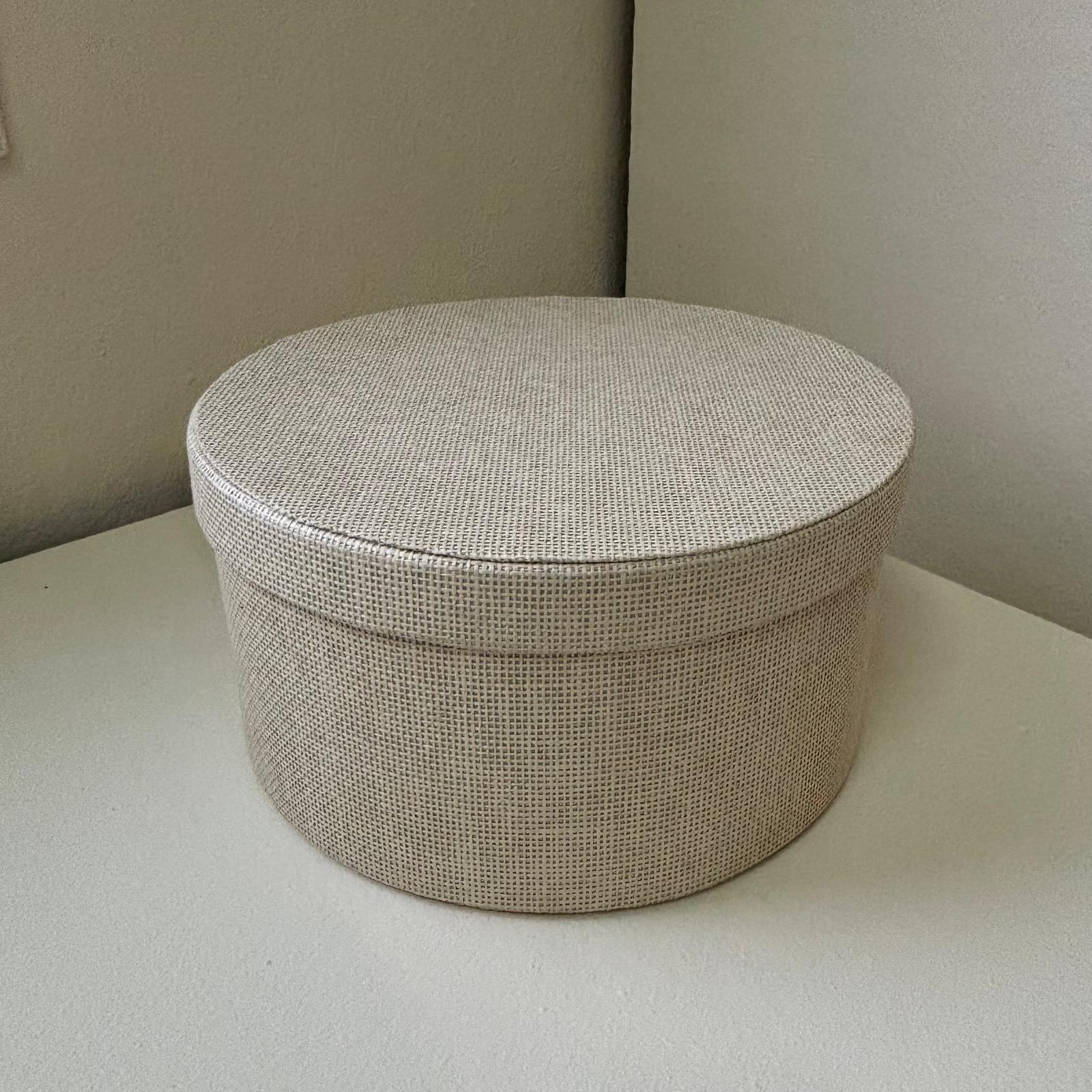 Cofanetto rotondo con coperchio, rivestito in tessuto beige chiaro a trama fitta, posizionato su una superficie chiara.
