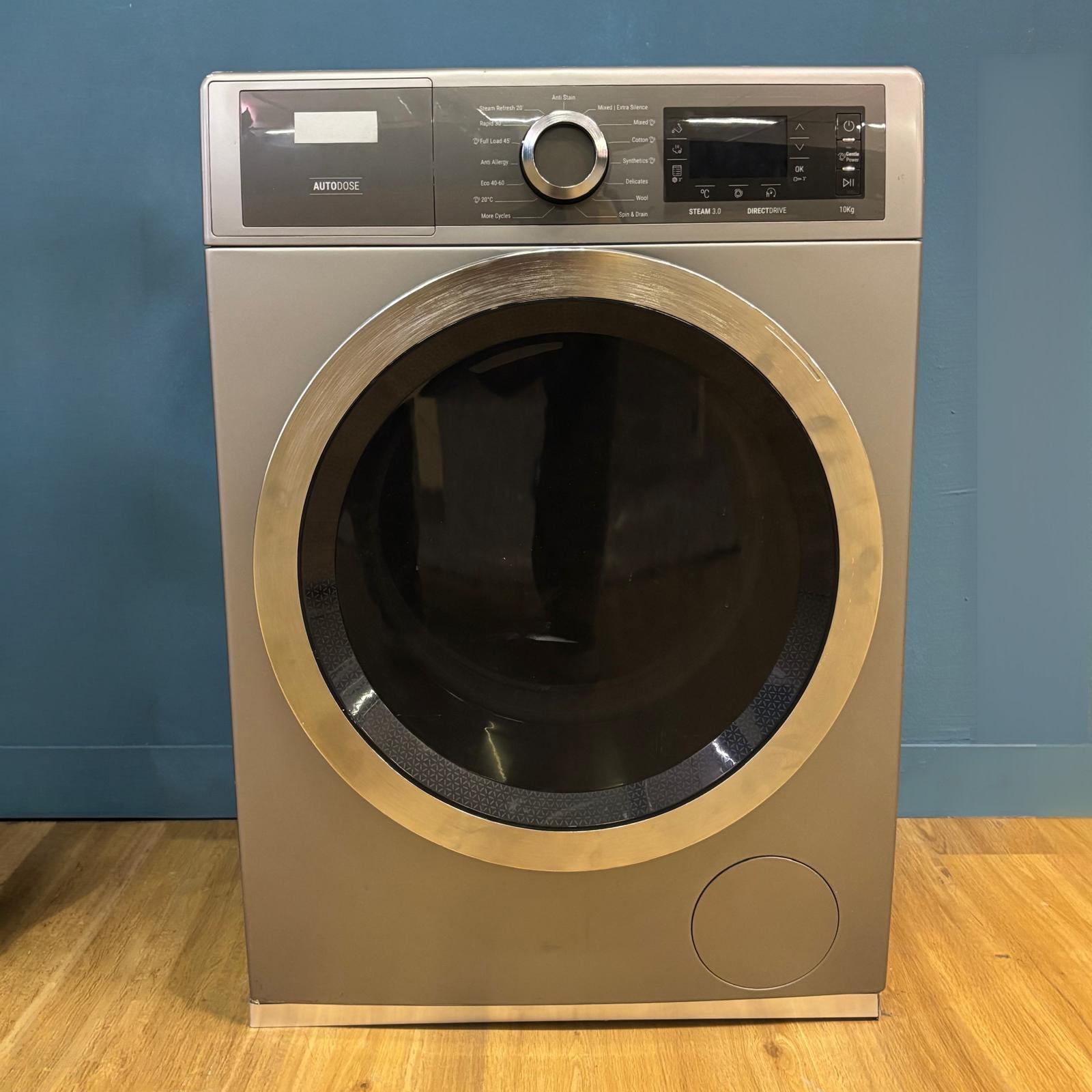 Lavatrice moderna grigia con oblò metallico e pannello di controllo digitale, posizionata su pavimento in legno davanti a parete blu.