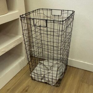Cesto portabiancheria rettangolare in filo metallico nero, con un panno chiaro all'interno, appoggiato su pavimento in legno chiaro.