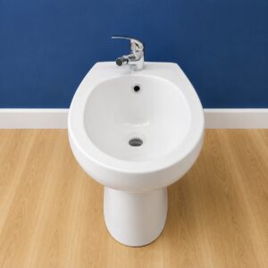 Bidet in ceramica bianca lucida con rubinetteria cromata, posizionato su pavimento in legno chiaro davanti a una parete blu.