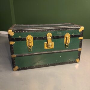 Baule vintage di colore verde scuro con fasce nere e borchie, angoli e chiusure frontali in ottone anticato, posizionato su un pavimento grigio davanti a una parete verde.