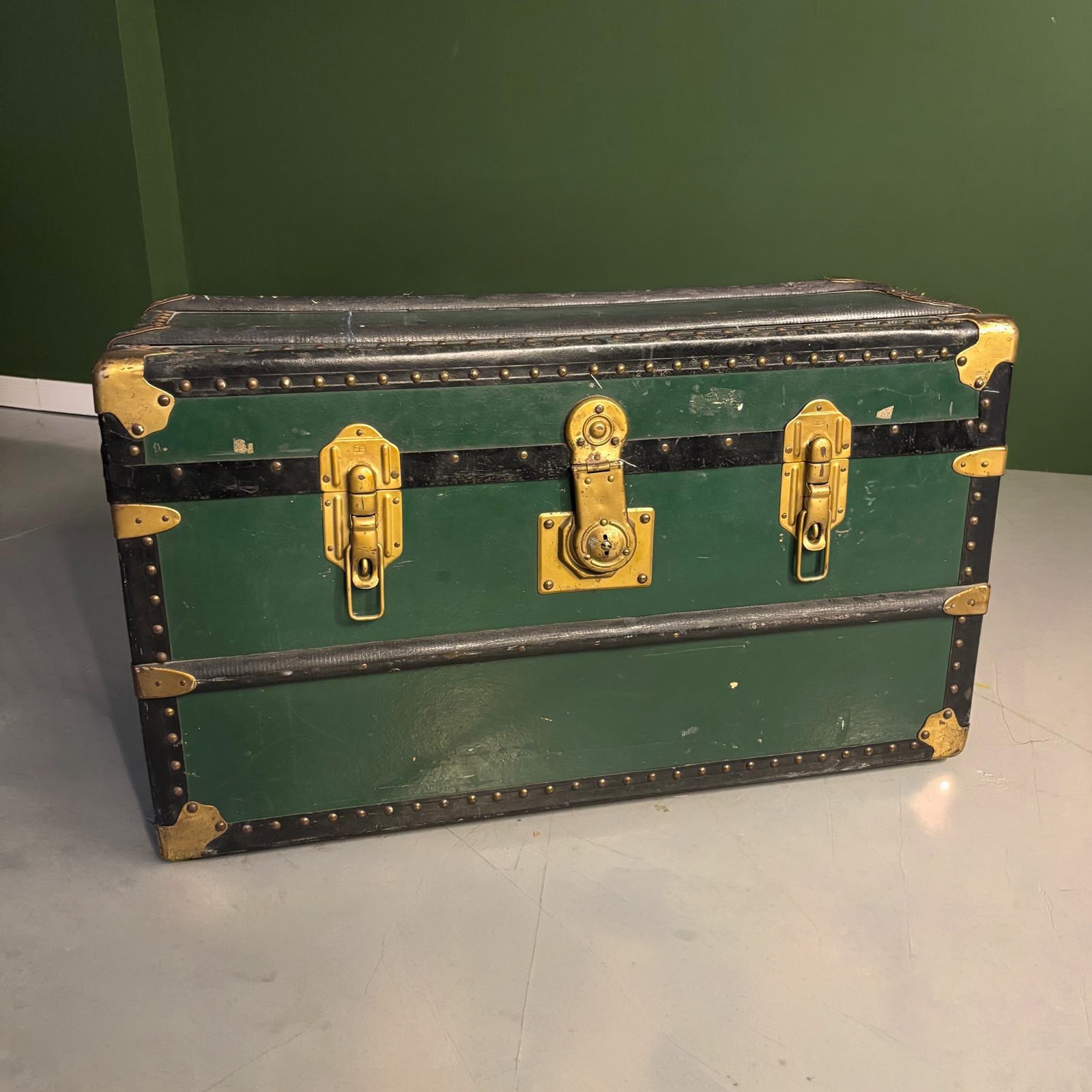 Baule vintage di colore verde scuro con fasce nere e borchie, angoli e chiusure frontali in ottone anticato, posizionato su un pavimento grigio davanti a una parete verde.