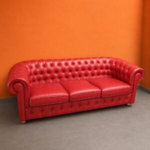Divano Chesterfield rosso a tre posti con rivestimento capitonné e braccioli arrotolati, su pavimento grigio con parete arancione.