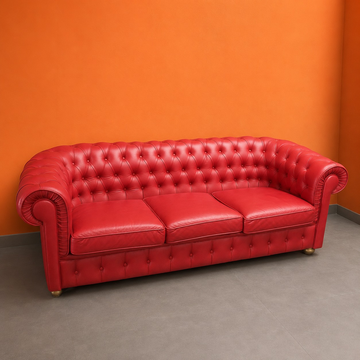 Divano Chesterfield rosso a tre posti con rivestimento capitonné e braccioli arrotolati, su pavimento grigio con parete arancione.