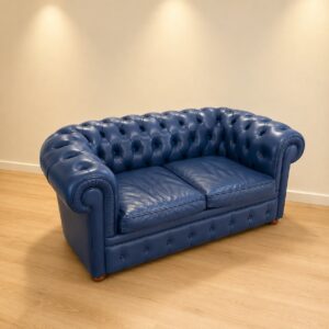 Divano Chesterfield a due posti in pelle blu, con bottonatura capitonné e braccioli arrotolati, su pavimento in legno chiaro.