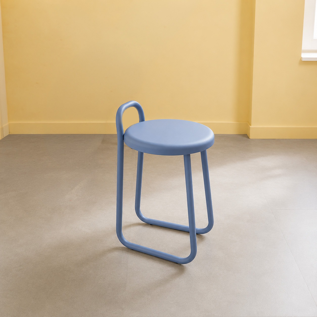 Sgabello monoblocco blu pastello con seduta rotonda e schienale curvo, su pavimento grigio chiaro con parete gialla.