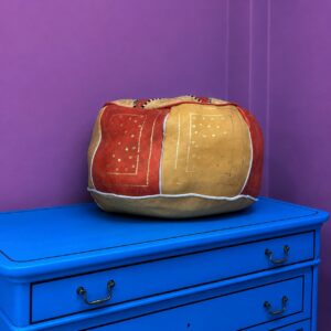 Pouf rotondo decorativo con design patchwork in tessuti rossi e oro, impreziosito da motivi dorati, su un mobile blu.
