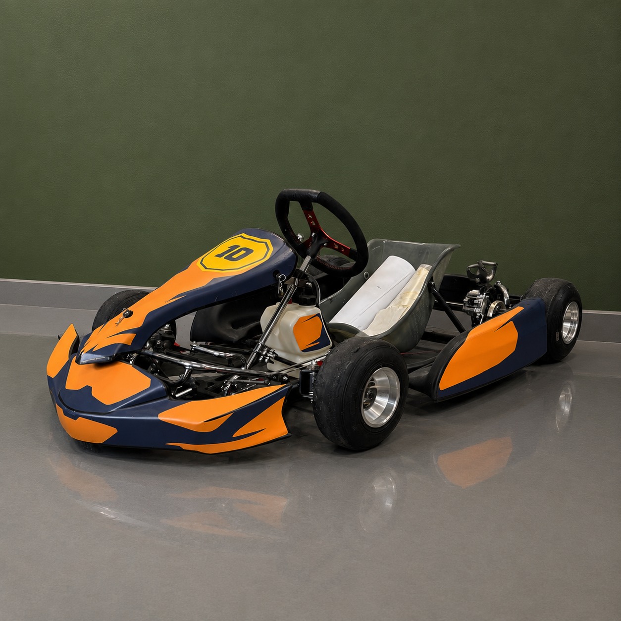 Kart da corsa arancione e blu con numero 10, posizionato su pavimento grigio davanti a una parete verde.
