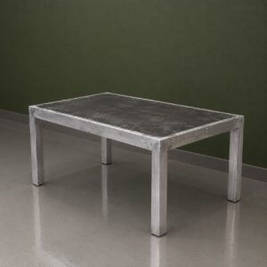 Tavolo rettangolare con struttura in alluminio spazzolato e piano scuro effetto cemento, su pavimento grigio con parete verde scuro.