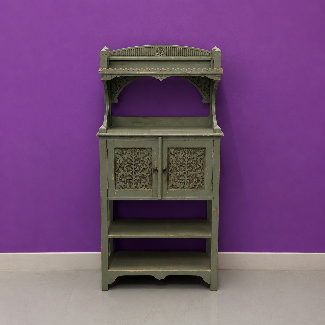 Mobiletto in legno verde-grigio anticato con ante intagliate e ripiani a vista, posto contro una parete viola.