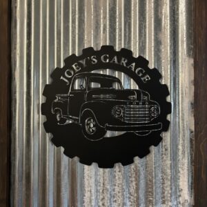 Insegna decorativa circolare in metallo nero a forma di ingranaggio con silhouette di pickup Ford e scritta "Joey's Garage", montata su lamiera ondulata.