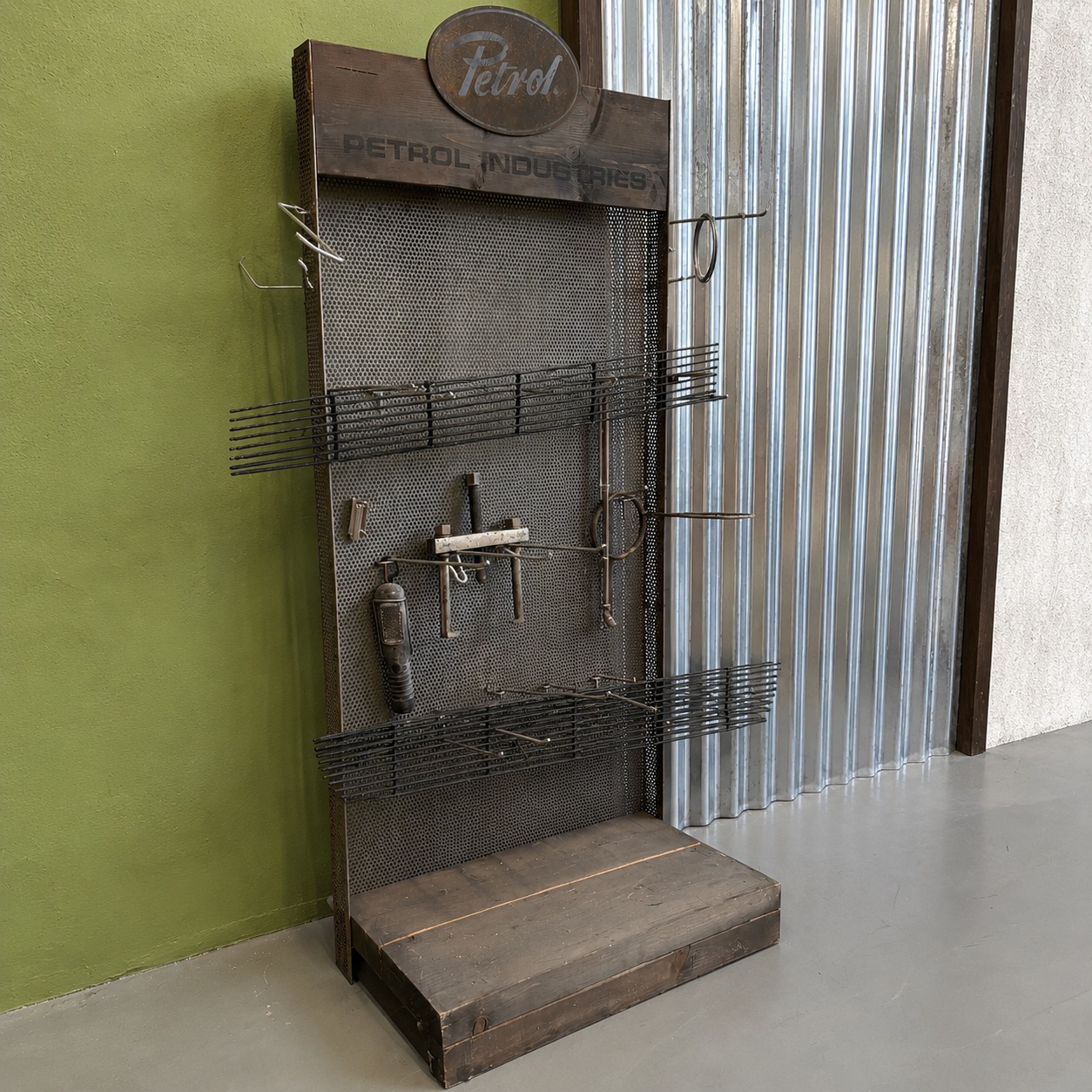Pannello espositivo industriale vintage in legno scuro e metallo forato con insegna 'Petrol Industries', ganci e mensole.