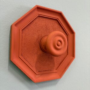 Maniglia decorativa ottagonale color terracotta con pomello centrale, montata su una parete chiara, con texture granulosa.