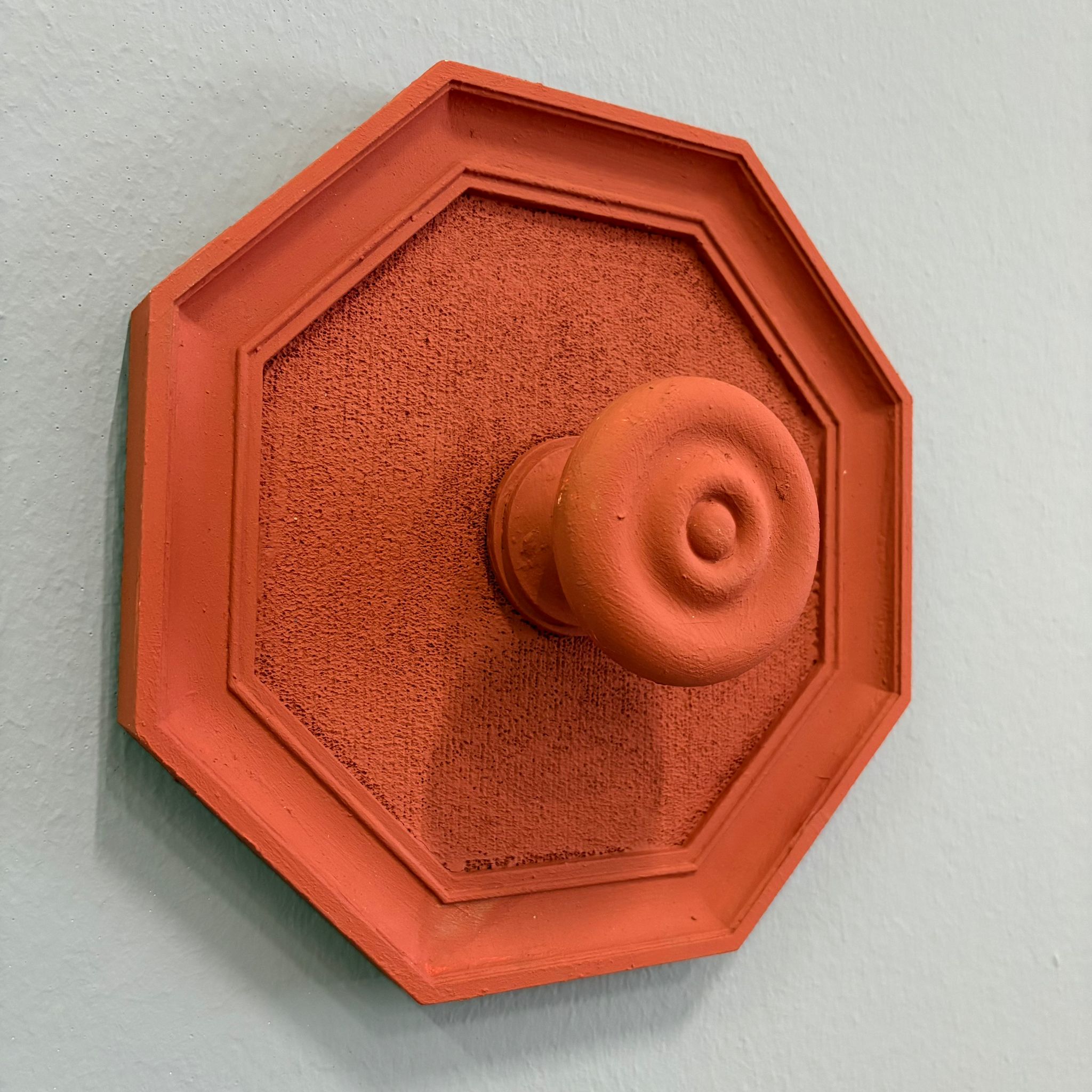 Maniglia decorativa ottagonale color terracotta con pomello centrale, montata su una parete chiara, con texture granulosa.