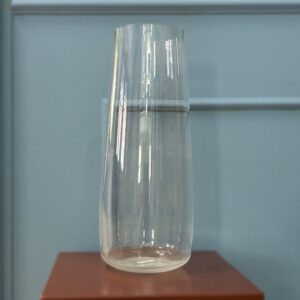 Caraffa in vetro trasparente, alta e cilindrica con leggera conicità, su una superficie marrone davanti a una parete grigio-azzurra.