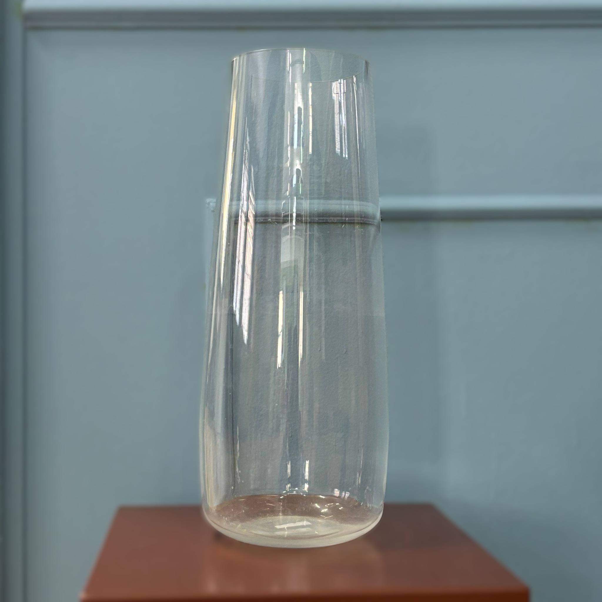 Caraffa in vetro trasparente, alta e cilindrica con leggera conicità, su una superficie marrone davanti a una parete grigio-azzurra.