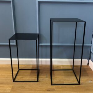 Due pedestali espositivi neri in metallo, di diverse altezze, con design minimalista, su pavimento in legno chiaro.