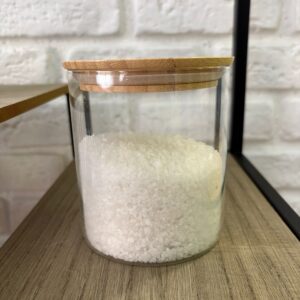 Barattolo cilindrico in vetro trasparente con coperchio in bambù, riempito di sale grosso bianco, posizionato su una mensola in legno.