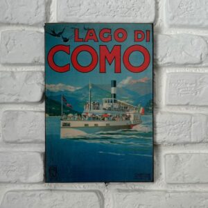 Poster vintage del Lago di Como con battello a vapore, montagne e rondini, appeso su muro di mattoni bianchi.