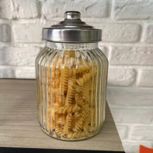 Barattolo di vetro trasparente con scanalature verticali, riempito di pasta fusilli, con coperchio metallico su ripiano in legno.
