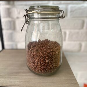 Vaso in vetro trasparente con chiusura ermetica a leva in metallo, riempito per due terzi con cereali soffiati marroni, posizionato su una mensola di legno chiaro con uno sfondo di parete in mattoni bianchi.