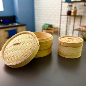 Due vaporiere in bambù di diverse dimensioni, una grande con coperchio sollevato e una piccola con coperchio, su un tavolo scuro in un ambiente cucina.