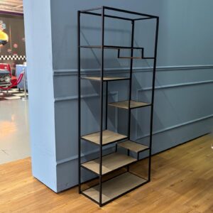 Scaffale alto con struttura in metallo nero e ripiani in legno chiaro disposti asimmetricamente, su pavimento in parquet.