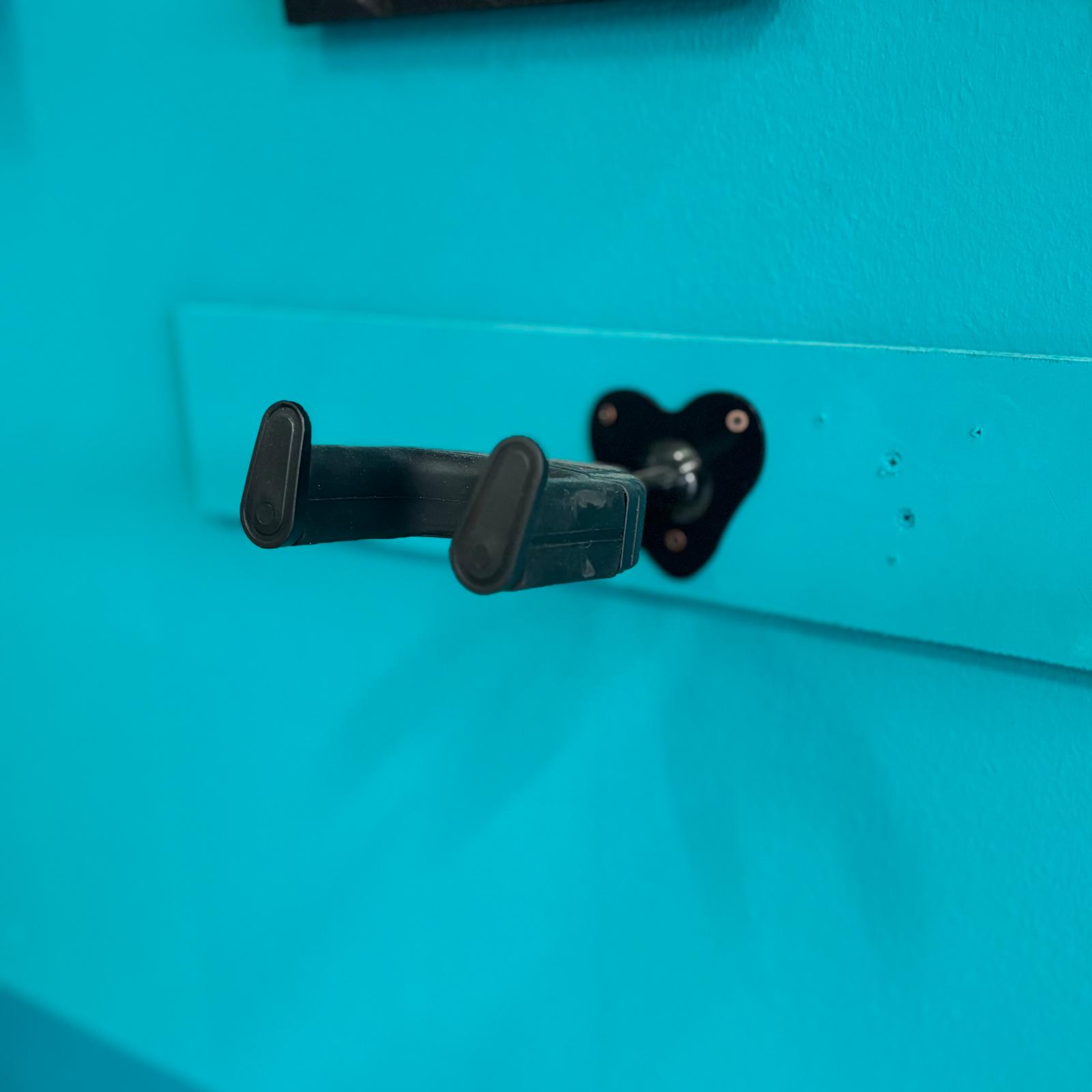 Gancio a muro nero con base a cuore e bracci imbottiti, montato su una tavola di legno azzurra, su una parete azzurra.