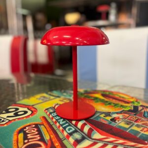 Lampada decorativa rossa lucida a forma di fungo, con base rotonda e stelo sottile, posizionata su un tappetino da tavolo colorato.