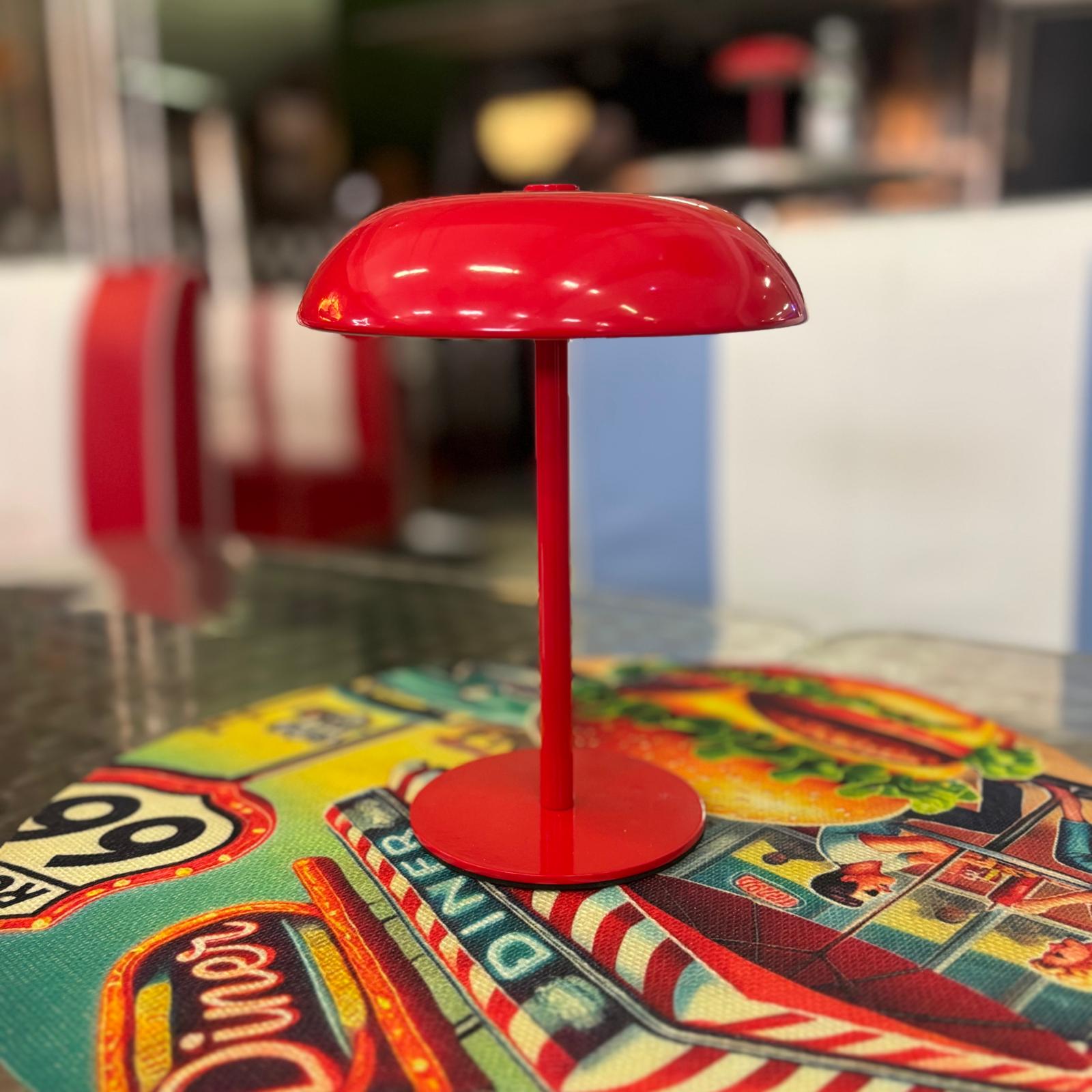 Lampada decorativa rossa lucida a forma di fungo, con base rotonda e stelo sottile, posizionata su un tappetino da tavolo colorato.
