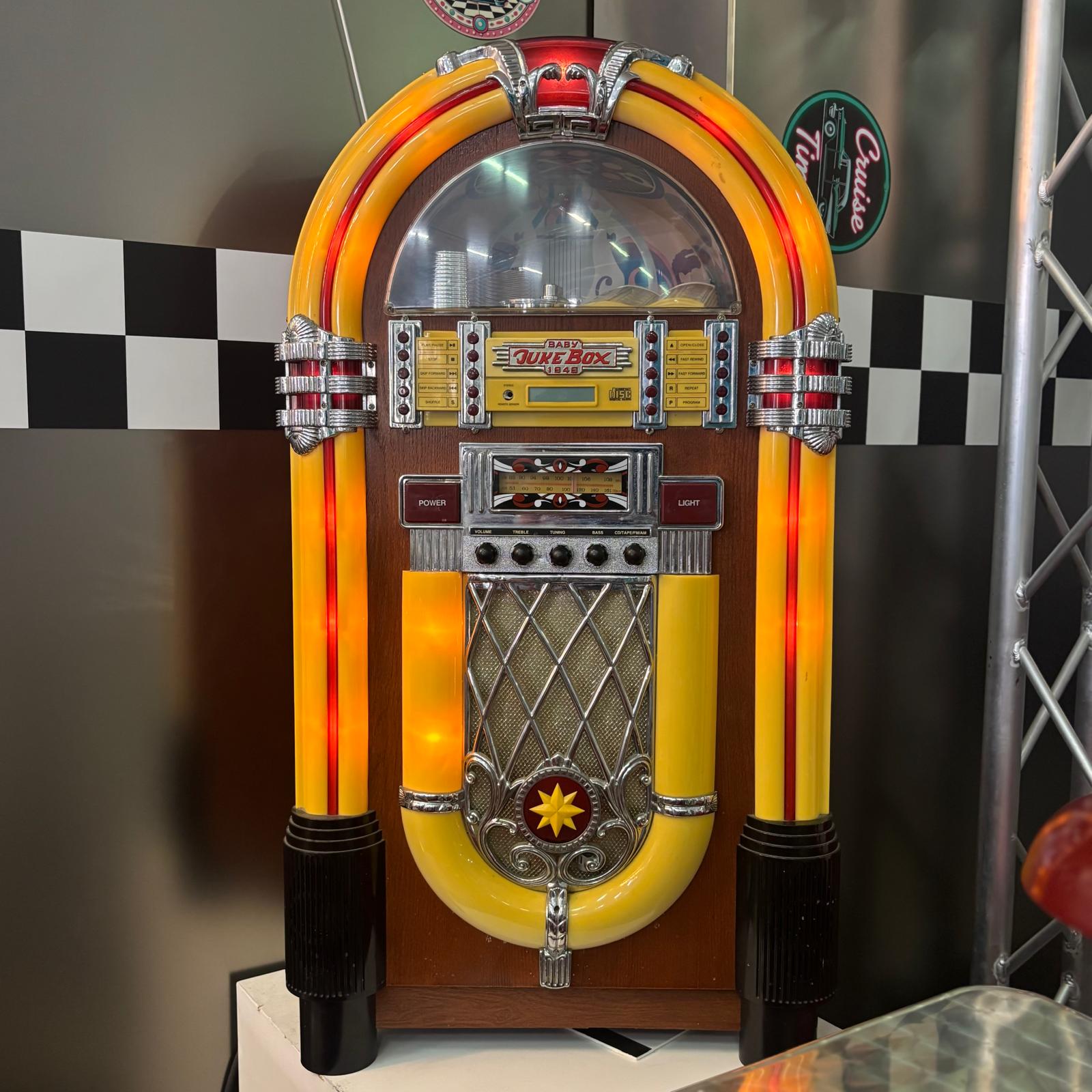 Jukebox decorativo vintage giallo e rosso con dettagli cromati e illuminazione interna, posizionato su sfondo a scacchi.