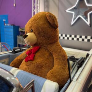 Orso gigante di peluche marrone con fiocco rosso seduto nel sedile posteriore di un'auto d'epoca in un hangar.