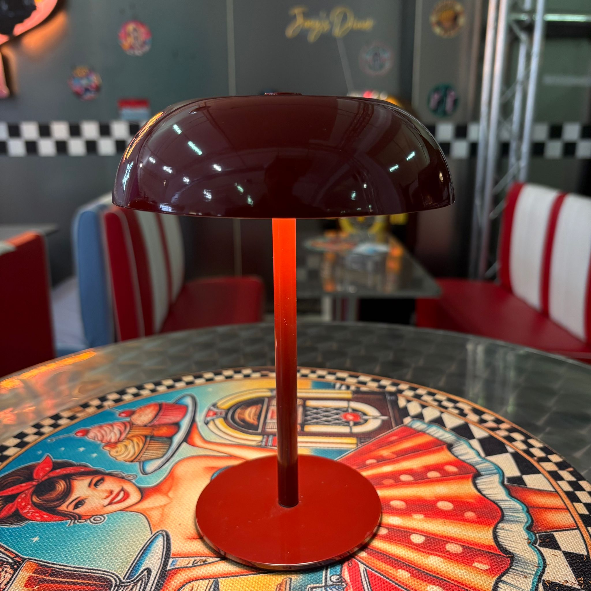 Lampada da tavolo rossa lucida a forma di fungo, accesa, su un tavolo con tovaglietta retro in un ambiente stile diner.