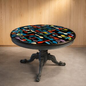 Tavolo rotondo con base in metallo scuro ornato e piano nero lucido decorato con posate stilizzate multicolori.