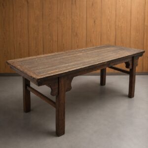 Tavolo rettangolare in legno massello scuro con finitura rustica e invecchiata, su sfondo di parete in legno e pavimento grigio.