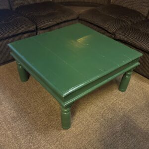 Tavolino quadrato in legno verniciato di verde scuro con gambe tornite, posizionato su un tappeto marrone chiaro.