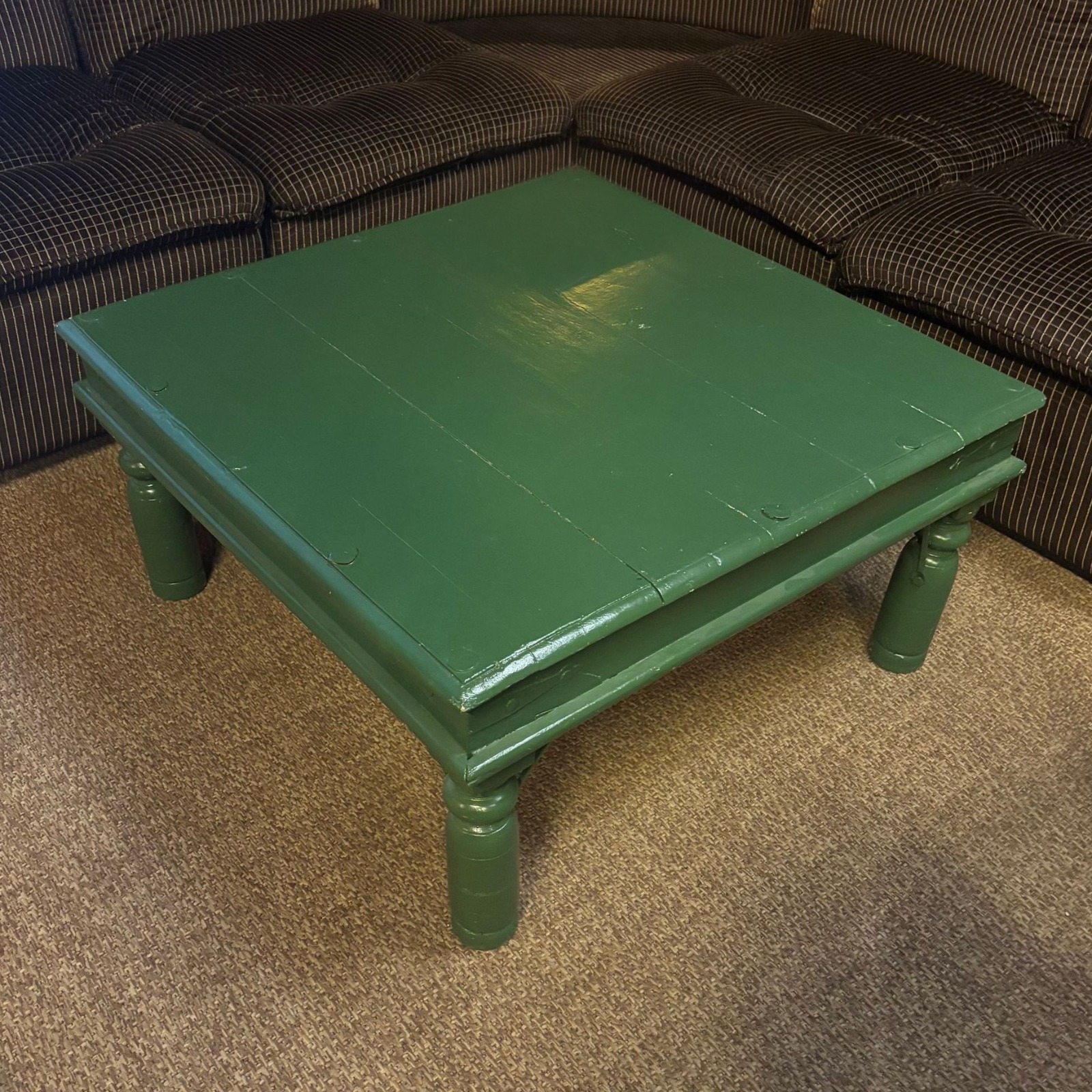 Tavolino quadrato in legno verniciato di verde scuro con gambe tornite, posizionato su un tappeto marrone chiaro.